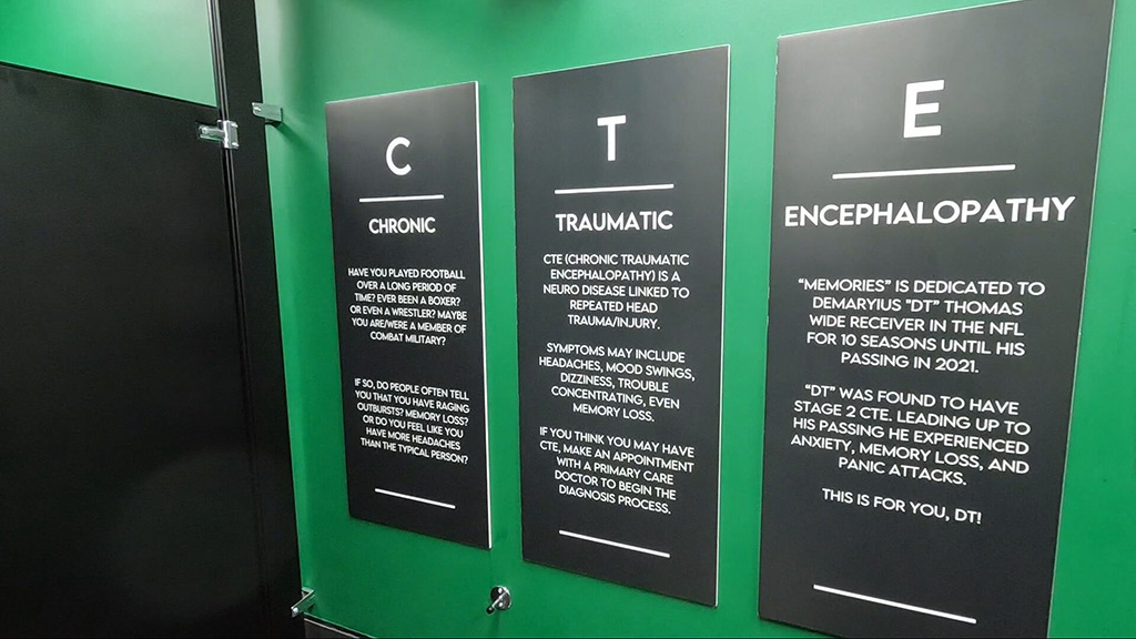 Three informational posters on a green wall at Memories Lounge. The posters discuss Chronic Traumatic Encephalopathy (CTE), detailing its symptoms and effects. One poster is dedicated to Demaryius “DT” Thomas, a former NFL player who passed away in 2021.